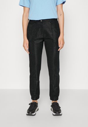 Personne portant un pantalon de jogging noir brillant sur mesure, une chemise bleu clair à manches courtes et des baskets athlétiques noires, debout devant un fond blanc.