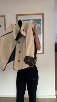 Bēšs mākslīgās kažokādas mētelis ar brūniem akcentiem un pogām, ar platu apkakli un redzamām šuvēm. Valkāts kopā ar melnām biksēm.