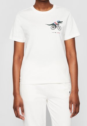 Osoba nosząca białą koszulkę z krótkim rękawem z małym nadrukiem aligatora w kasku na górnej lewej części klatki piersiowej, w połączeniu z białymi spodniami.