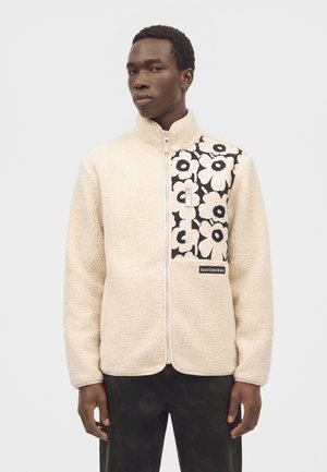 Joven con chaqueta de forro polar beige con estampado floral en blanco y negro en el pecho y cremallera frontal.