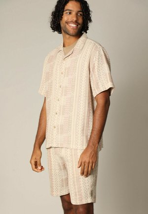 Beige set van een kortarmige shirt en shorts met een getextureerd geometrisch patroon. Knoopsluiting op het shirt, ontspannen pasvorm, lichte stof.