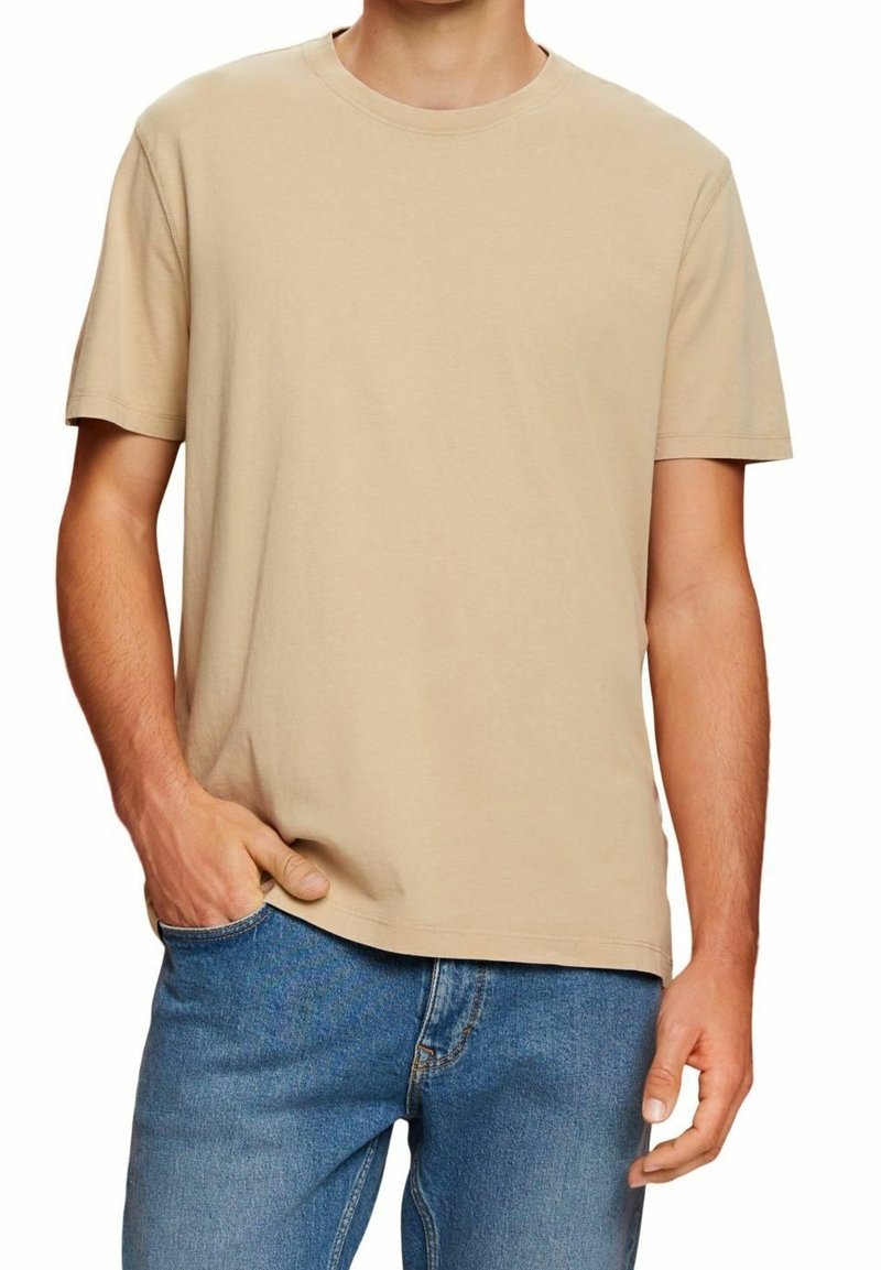 T-shirt en coton beige avec un col rond et des manches courtes, porté par une personne vêtue de jeans bleus, mettant en valeur une coupe décontractée et une texture lisse.