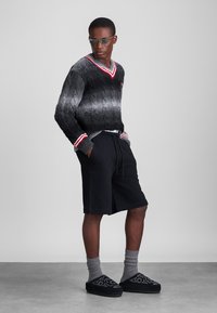 Zwart en grijs gebreid trui met een V-hals en rode accenten, gecombineerd met zwarte shorts, grijze sokken en zwart met patroon instappers.
