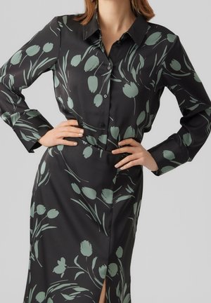 Vrouw draagt een zwarte jurk met knopen met groene bloemenprint, lange mouwen, kraag en voorste split, handen rustend op haar taille.