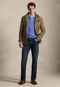 Jeune homme portant un blazer en tweed marron, un pull bleu torsadé sur une chemise blanche, un jean foncé, des mocassins marron et une montre-bracelet, debout avec une main dans la poche.