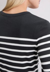 Blond person iført en svart og hvit stripete langermet topp, vist fra bak og siden med en enkel lys bakgrunn.