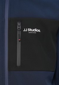 Marineblå jakke med en sort brystlomme. Har lynlås og hvidt logo "JJ Studios. Jack & Jones" på lommen.