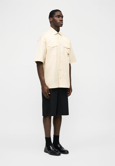 Homme portant une chemise beige à manches courtes avec boutons, un short noir jusqu'aux genoux, des chaussettes noires et des chaussures en cuir noires, debout devant un fond blanc.