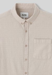 Chemise boutonnée beige clair avec un motif texturé, un col et une poche avant. Présente des boutons assortis le long de la patte de boutonnage.