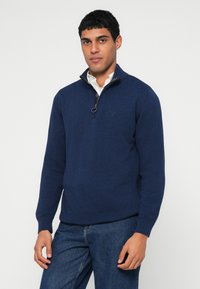 Camisola de fecho azul marinho com gola alta, feita de tecido macio, com um logótipo subtil no peito e punhos e bainha canelados.