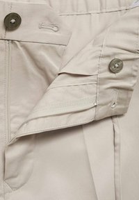 Beige fabric waistband with button closure, zip pocket, and flat-textured surface. Features stitching and side panel design for fit.