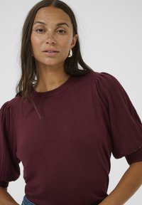 Burgundy stickad topp med ribbade puffärmar, rund halsringning och en slät yta. Minimalistisk design, inga synliga mönster eller hårdvara.