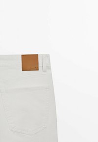Jeans in denim bianco con una cucitura in pelle marrone in vita. Le caratteristiche includono una singola tasca posteriore e dettagli di cucitura sottili.