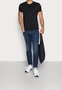 T-shirt noir à manches courtes, jeans fitted bleu foncé, baskets blanches avec des accents noirs et rouges. Sac fourre-tout noir à la main. Fond neutre.