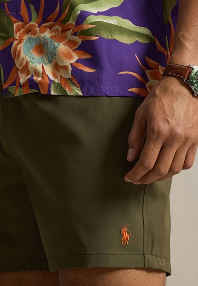 Κοντινό πλάνο ενός ατόμου που φοράει ελιά πράσινο σορτς με πορτοκαλί λογότυπο αλόγου και μοβ πουκάμισο με λουλούδια, με ρολόι χειρός στο αριστερό χέρι ορατό.