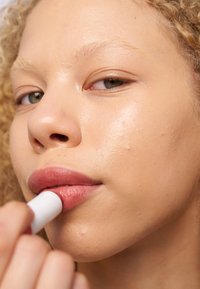 Un bálsamo labial se aplica a unos labios llenos y suavemente coloreados. La piel se ve suave con un ligero brillo, y el fondo es neutral y difuminado.