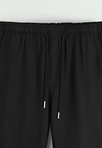 Pantalons noirs avec une taille élastique et des cordons de serrage noués avec des embouts en métal, montrés de la taille jusqu'à mi-cuisse.