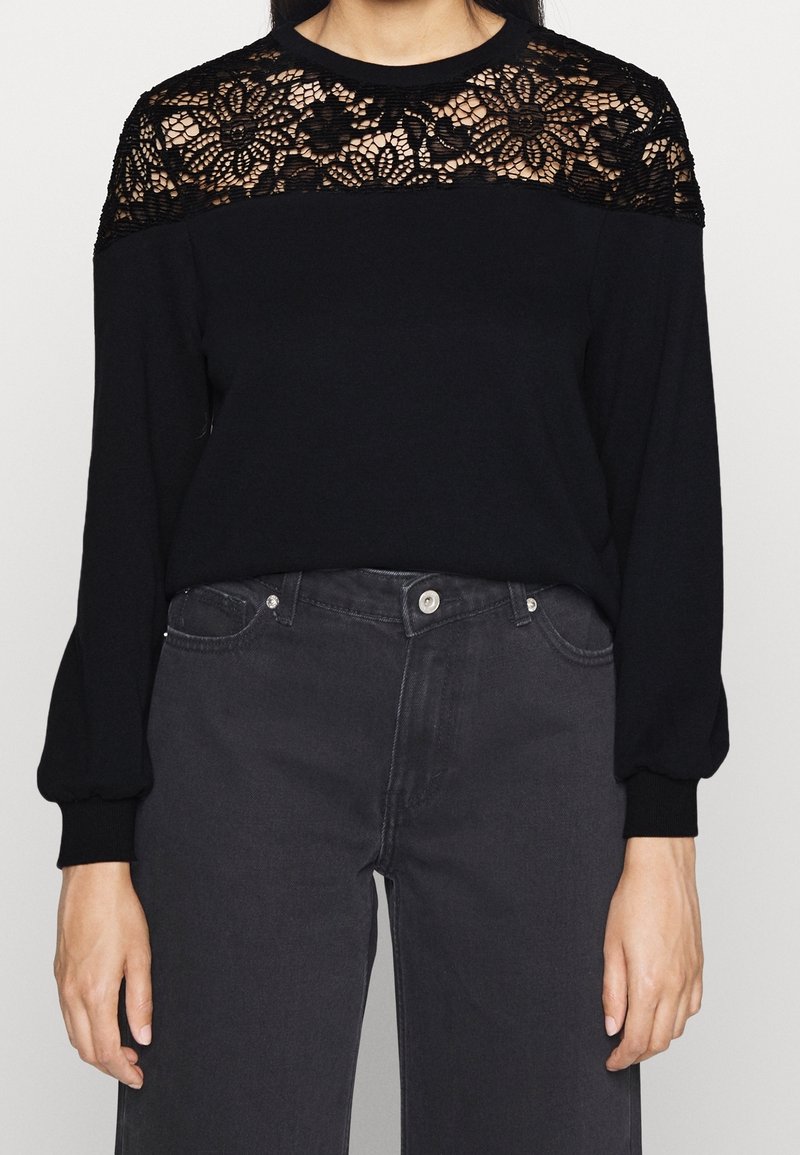 Femme portant un haut noir à manches longues avec un détail en dentelle florale sur les épaules et le haut de la poitrine, associé à un jean gris foncé taille haute.
