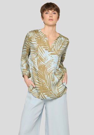 Woman with short brown hair wearing a loose brown and light blue palm leaf patterned blouse and light blue wide-leg pants with hands in pockets.
