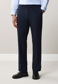 Uomo con pantaloni da abito blu scuro, camicia azzurro chiaro e scarpe nere, in piedi davanti a una parete liscia e chiara.