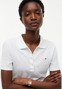 Femme aux cheveux courts et bouclés portant un polo blanc avec un petit logo et une montre-bracelet en or, bras croisés.
