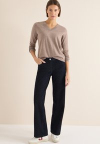 Vaaleanruskea v-kaula-aukkoinen neule yhdistettynä tummansinisiiin levenevälahkeisiin farkkuihin, joissa on hienovaraiset vinorivitomukset, ja valkoisiin lenkkareihin.