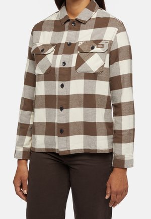 Kobieta w brązowo-białej koszuli w kratę na guziki z kieszeniami na piersi, w połączeniu z ciemnobrązowymi spodniami, widoczna od szyi do bioder.