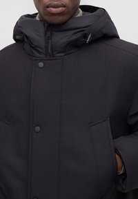 Homme portant une veste d'hiver matelassée noire avec un col montant, une capuche, une fermeture éclair, des boutons-pression et les mains dans les poches avant.