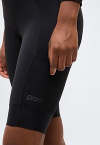 Κοντινή λήψη ενός ατόμου που φοράει μαύρο εφαρμοστό σορτς ποδηλασίας με δίχτυ στη πλαϊνή τσέπη και λογότυπο "poc", το χέρι χαλαρό δίπλα στο μηρό.