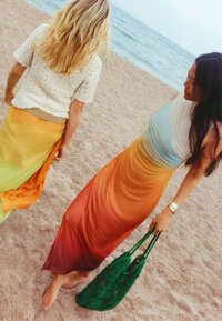 Zwei Frauen am Strand, eine in einem orangefarbigen Maxikleid mit Farbverlauf, die andere in einem gelb-orangefarbenen Rock und einem gestrickten cremefarbenen Oberteil. Beide barfuß.