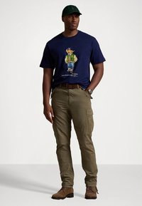 Navy t-shirt featuring a colorful bear graphic, olive green cargo pants, and brown boots. The outfit is completed with a dark green cap.