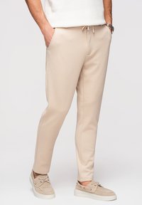 Homme portant un pantalon beige resserré avec des poches, associé à des chaussures bateau beige et un haut blanc texturé, debout les mains dans les poches.