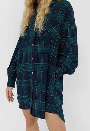 Femme portant une chemise oversize à carreaux vert foncé et bleu marine, manches longues, debout devant un fond clair uni.