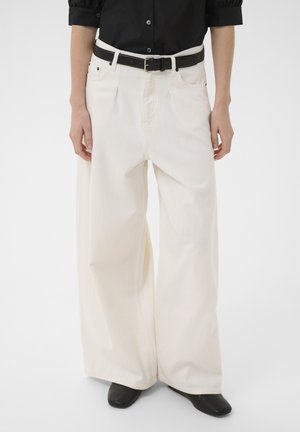 Personne portant un pantalon blanc à jambes larges, une ceinture noire, une chemise noire boutonnée et des chaussures noires à bout carré sur un fond uni.