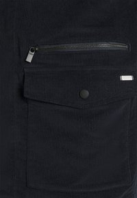 Schwarze Cordjacke mit einer Reißverschlusstasche und einer Klappentasche mit Druckknopfverschluss. Strukturierter Stoff mit vertikalen Rillen.