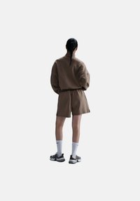 Conjunto de ropa de estar en casa marrón que incluye una sudadera con cremallera de corte holgado y pantalones cortos a juego. La prenda tiene una tela texturizada y se combina con calcetines blancos y zapatillas.