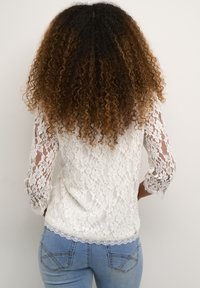 Ženska s kodrastimi temno rjavimi lasmi, oblečena v belo čipkasto majico z dolgimi rokavi in svetlo modre kavbojke, stoji obrnjena stran proti enobarvnemu ozadju.