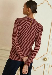 Dugog rukav s topom u tamnocrvenoj i krem prugastoj kombinaciji s keyhole detaljem na leđima i volanima na rukavima, uparen s tamnim hlačama. Glatka tkanina, prilagođen dizajn.
