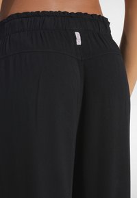 Mustat korkeavyötäröiset housut pehmeästä kankaasta, joissa on kerätty resorivyötärö ja takana pieni merkki brändin nimellä.