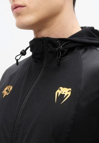 Μαύρο μπουφάν με φερμουάρ και κουκούλα, με χρυσά ζωικά λογότυπα στο στήθος. Το ύφασμα είναι υφασμένο με υφή, με λεπτομέρειες από διχτυωτό ύφασμα στους ώμους.