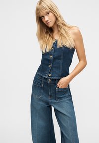 Femme blonde portant un haut en jean sans bretelles boutonné et un pantalon large en jean, une main dans la poche, sur un fond uni.