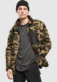 Rohelise ja beežikae kamuflaažiga fliisjope, millel on lukuga must rinnatasku ja kõrge kraega disain. Tekstuuriga ja soe.