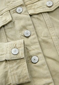 Beige denim overhemdsectie met zilveren drukknoopjes en zichtbare stiksels. De stof heeft een gestructureerde uitstraling met een lichte glans.