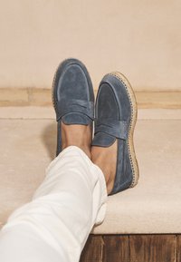 Mocassins en daim bleu avec un motif cousu, présentant une texture douce et une semelle en jute naturel. Portés avec un pantalon blanc court.