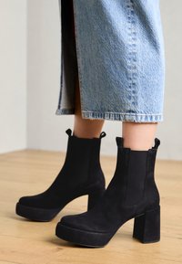 Bottines en cuir suédé noir avec un large talon bloc et une plateforme, dotées de panneaux élastiques sur les côtés et de languettes pour faciliter l'enfilage.