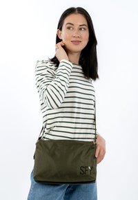 Oliwkowa torba na ramię z teksturowanej tkaniny, z regulowanym paskiem i widocznym logo. Modelka nosi pasiastą bluzkę z długim rękawem i dżinsy.