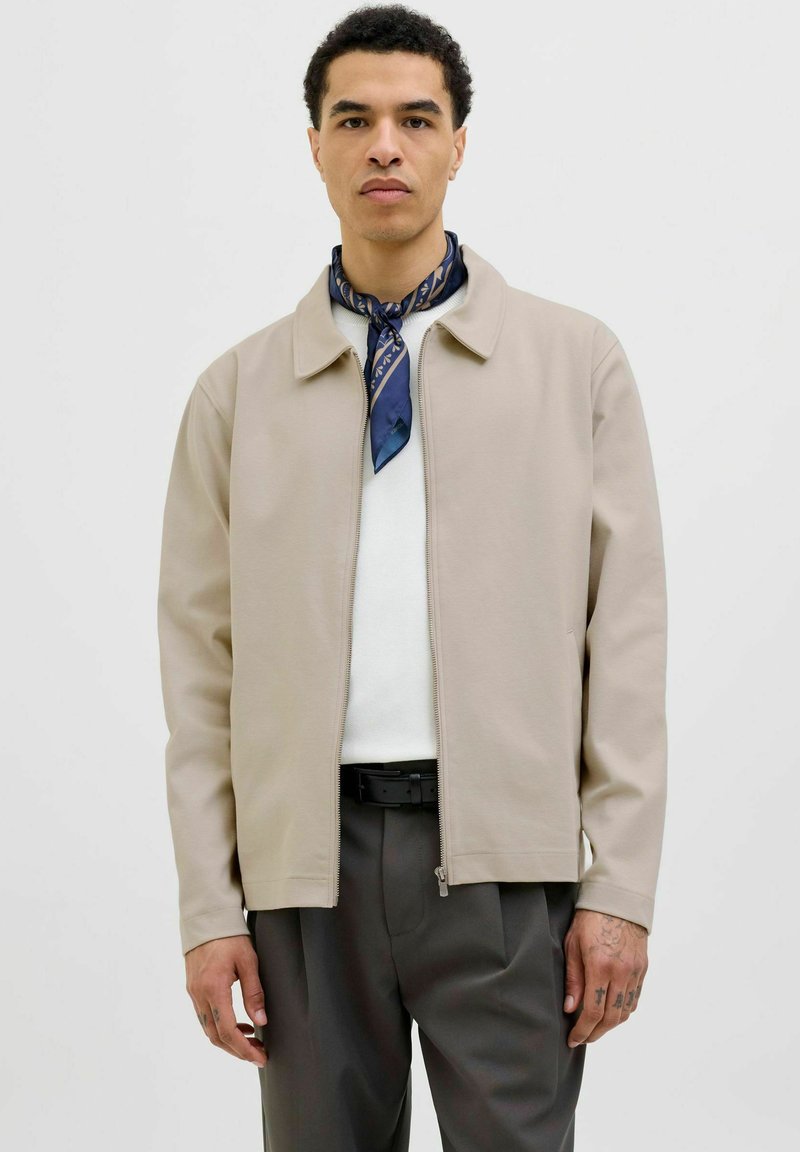 Beige ritsjack met een kraag, gladde textuur en zijzakken. Gemaakt over een witte trui en gecombineerd met grijze broeken.