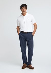 Witte poloshirt met een klein zwart logo, gecombineerd met marineblauwe chinos en bruine bootschoenen met oranje veters, staand tegen een lichte achtergrond.