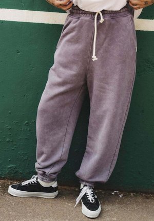 Osoba w luźnych fioletowych spodniach dresowych z białym troczkiem i czarno-białych trampkach, stojąca na tle zielonej ściany z białym paskiem.