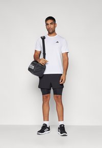 Weißes T-Shirt mit schwarzem Logo, schwarze Shorts über schwarzen Kompressionsshorts, schwarze Sneakers und eine schwarze Sporttasche mit weißem Logo.